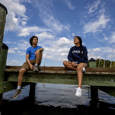 Two students sitting on a dock by the river with blue sky and white clouds.