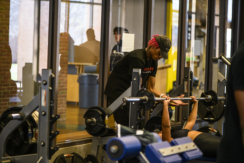 In the weight room of the Michael P. O'Brian Athletics Recreation Center a student spots another student on a weights bench