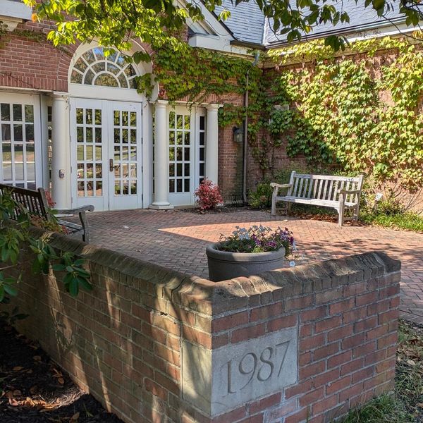 A brick building entrance with tall white doors and ivy-covered walls. A bench sits on the red brick patio near a flower planter. The year "1987" is engraved on a corner brick. Green leaves hang from a nearby tree, casting shadows on the scene.