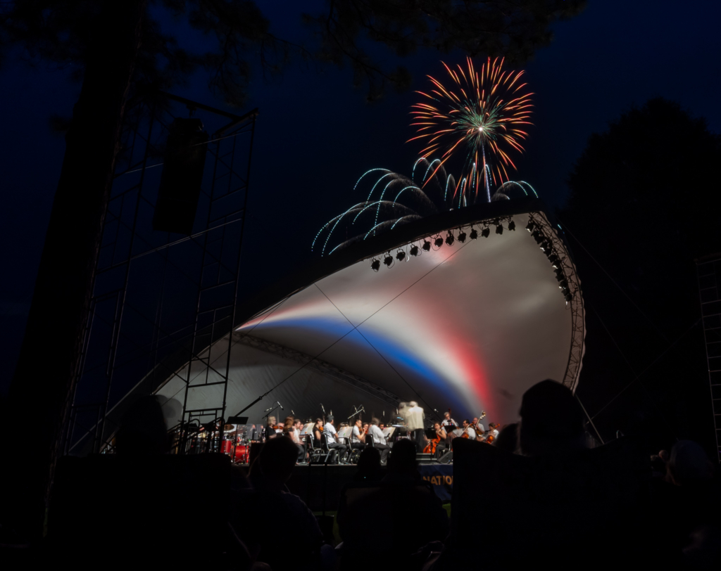 Fireworks bursting over orchestra playing under outdoor concert shell.